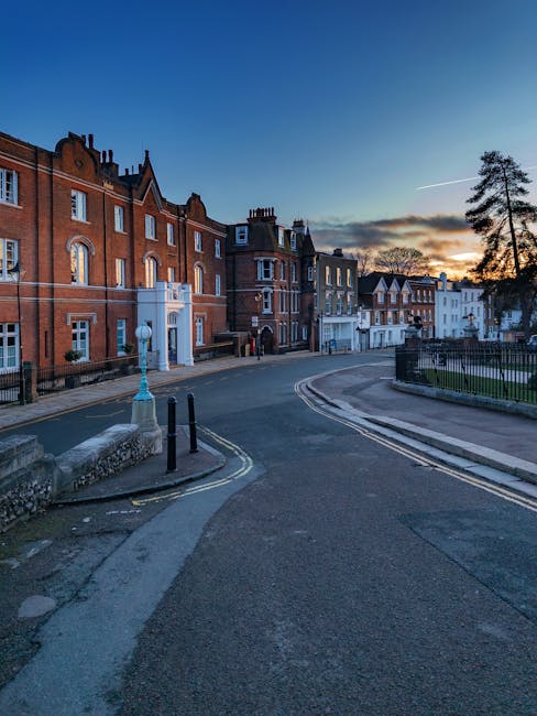 A paved street in Harrow, featuring a large historic church with a tall, pointed spire on the right side of the image, and red brick buildings with pitched roofs and chimneys lining the left side. Sev